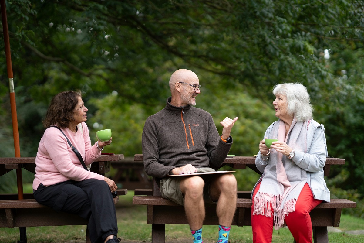 stock photo of three older adults drinking tea and talking while sitting on a park bench